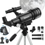 A black Qniuco telescope with multiple lenses and accessories is set on a tripod. A smartphone adapter displays a moon image, and a large moon is visible in the background.