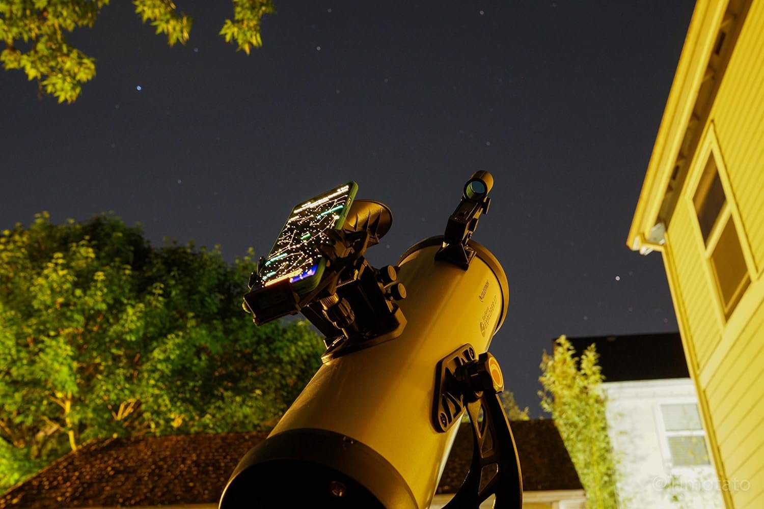 Telescope with an attached smartphone under a clear night sky, surrounded by trees and a building.