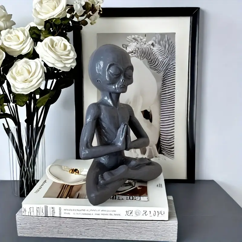 Gray alien figurine in a meditative pose sits on stacked books. Behind it is a black-framed photo of a zebra's ear, and a vase of white roses on the left side.
