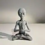 A small, gray alien statue is seated in a meditative pose with its eyes closed and hands pressed together, placed against a neutral background.