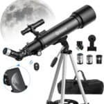 A black refractor telescope on a tripod with multiple eyepieces, a smartphone adapter, remote control, carry bag, and the moon visible in the background.