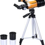 A compact orange and black telescope mounted on a silver tripod, with two additional eyepieces placed beside it.