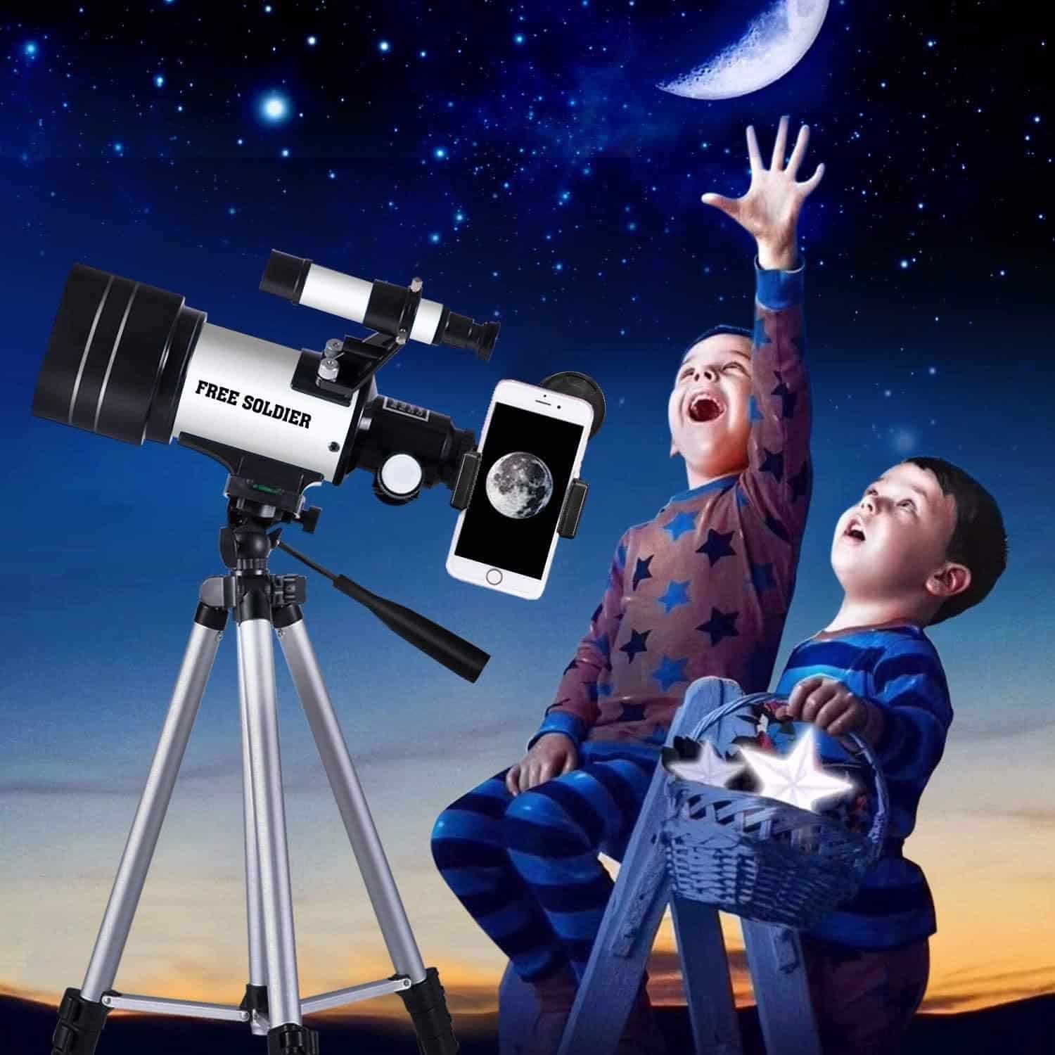 Two children in pajamas sit outdoors at dusk with a telescope and smartphone, looking excitedly at the crescent moon in the starry sky.