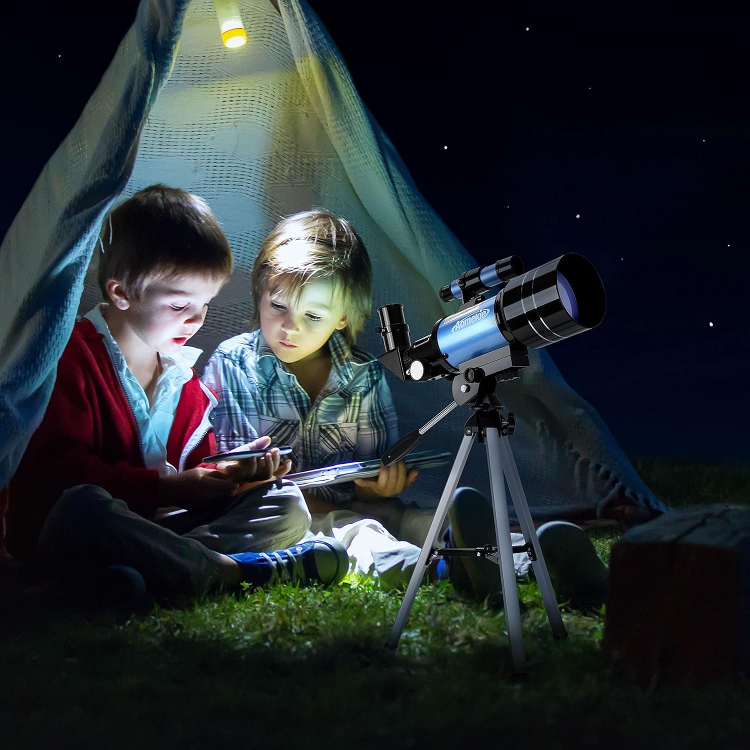 Two children sit inside a tent at night, illuminated by a light, looking at a book or device, with a telescope set up outside under the starry sky.