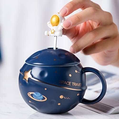 A hand lifts a lid topped with an astronaut figure from a dark blue, planet-themed mug with "SPACE TRAVEL" and space illustrations on it.