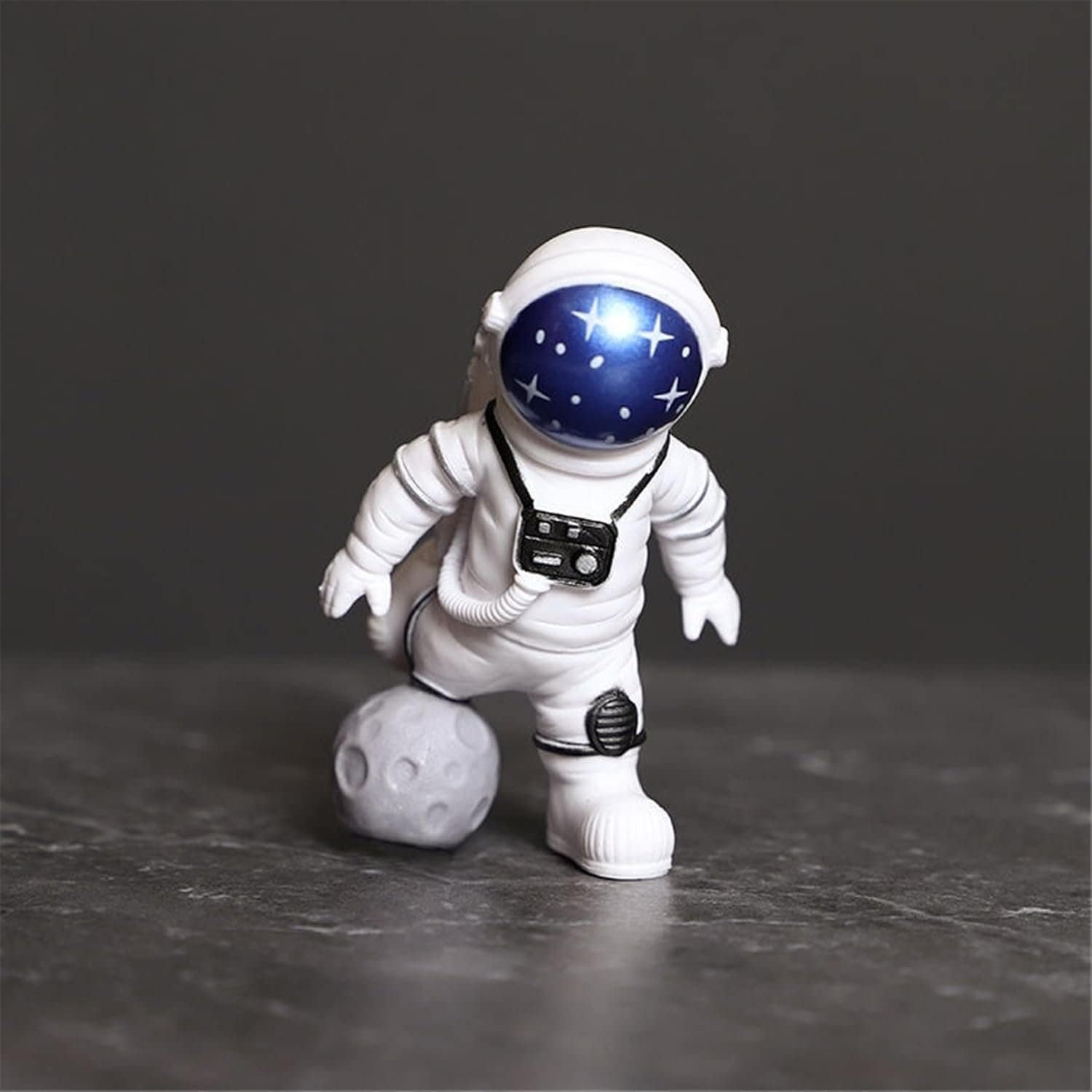 A small astronaut figurine in a white suit stands on a gray surface with one foot on a textured gray sphere resembling a moon or rock.