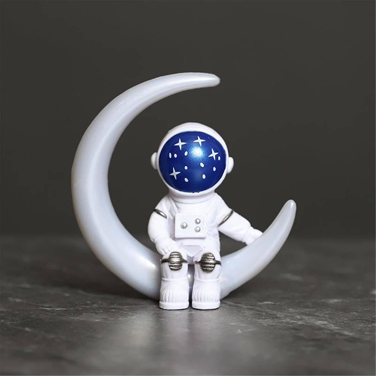 A small astronaut figurine with a starry night sky helmet sits on a crescent moon against a dark background.