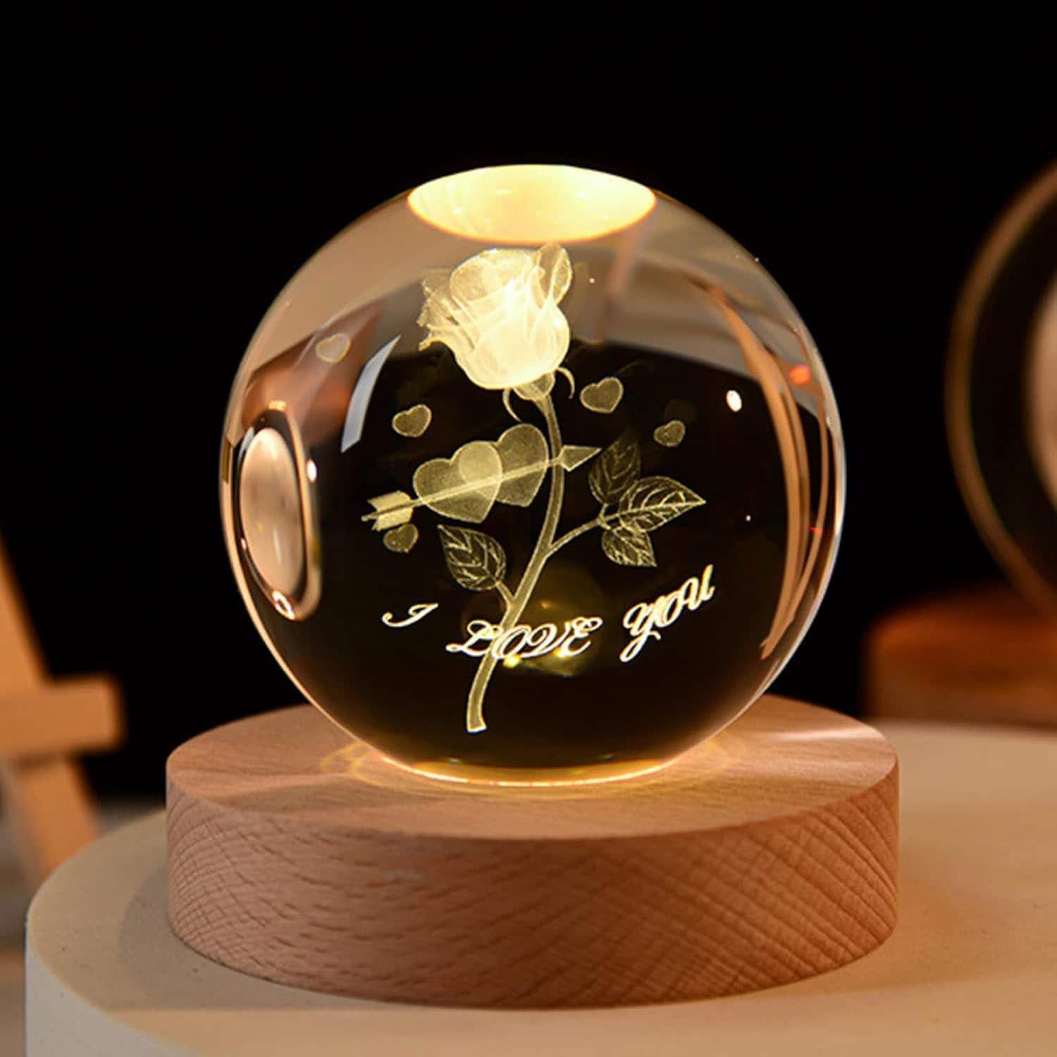 A crystal ball with an engraved rose, hearts, and the words "I love you," displayed on a wooden base and illuminated from above.