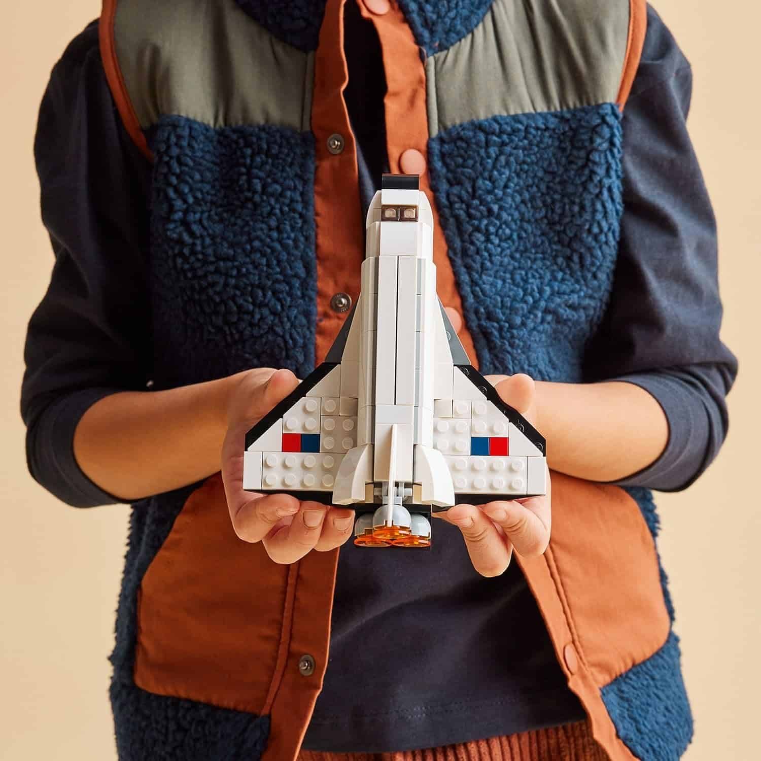 A child holds a LEGO space shuttle model with both hands, wearing a blue and orange vest over a navy shirt.