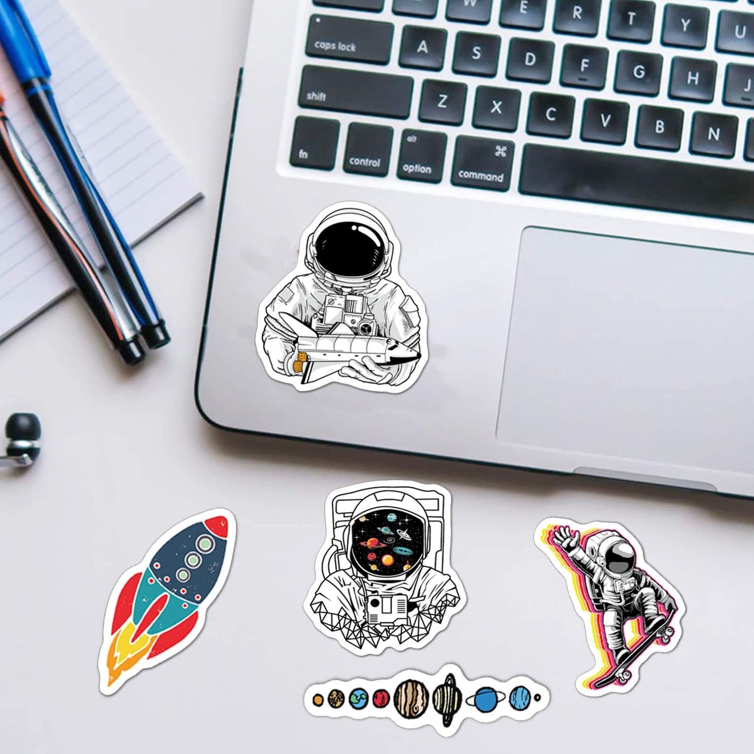 A laptop on a desk with pens, a notepad, and several space-themed stickers, including astronauts, a rocket, and planets, placed nearby and on the laptop.