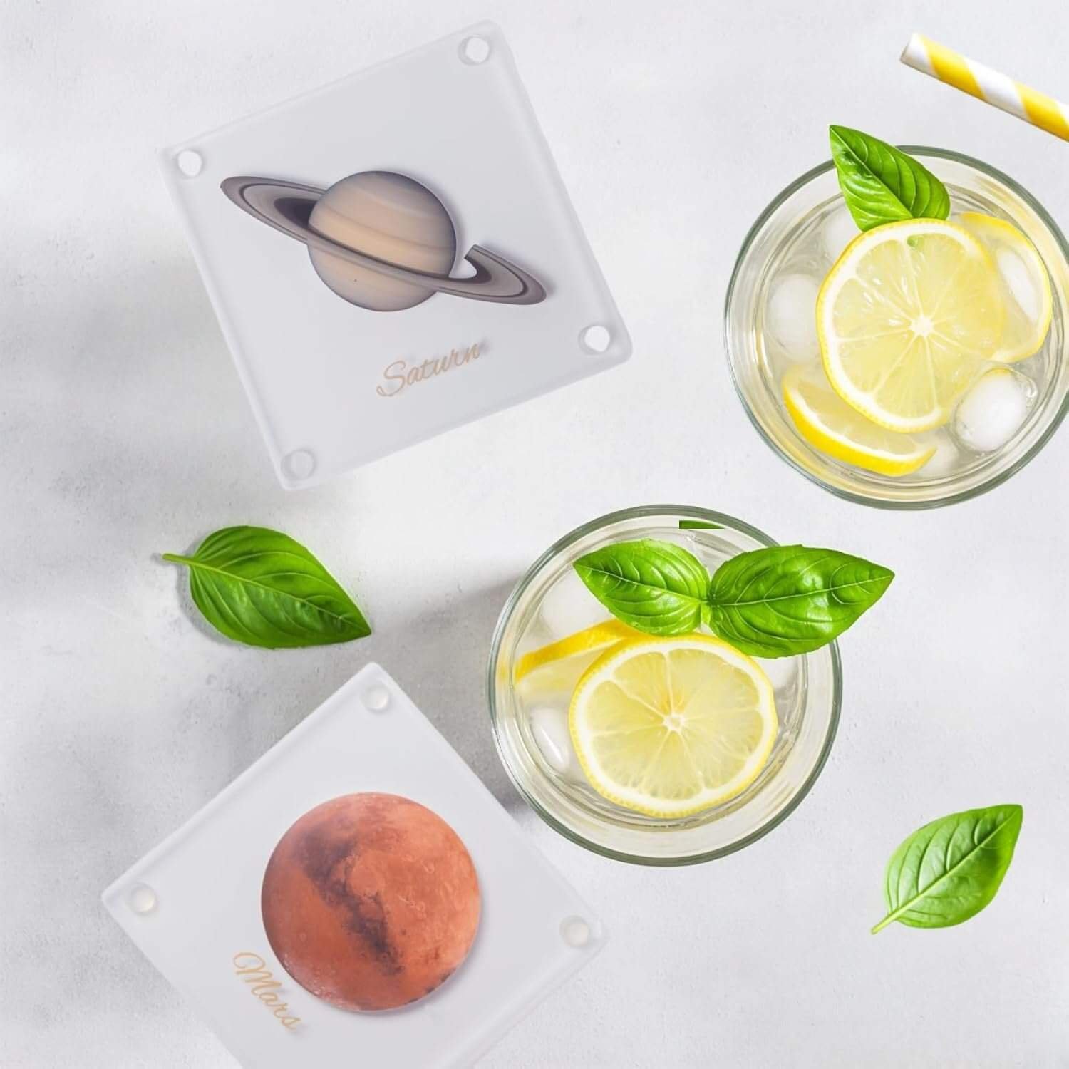 Two glasses of lemon water with basil leaves are next to two square coasters featuring images and names of Saturn and Mars on a white surface.