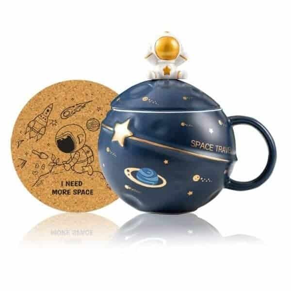 A blue space-themed mug with planets and stars, a lid topped with a small astronaut figure, and a cork coaster featuring space illustrations and the phrase "I NEED MORE SPACE.