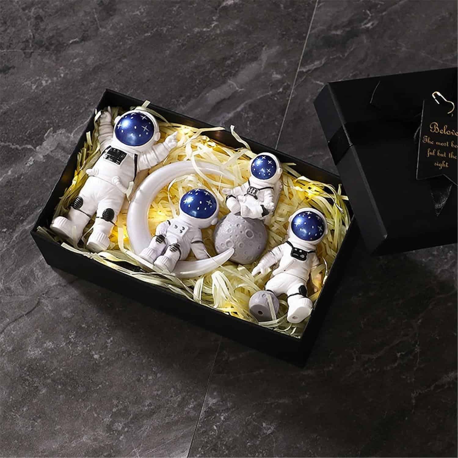 A black gift box contains four astronaut figurines, a crescent moon, and a small gray planet, all arranged on yellow shredded paper.