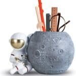 A moon-themed pen holder with a small astronaut figure beside it, containing pens, pencils, a ruler, and eyeglasses.