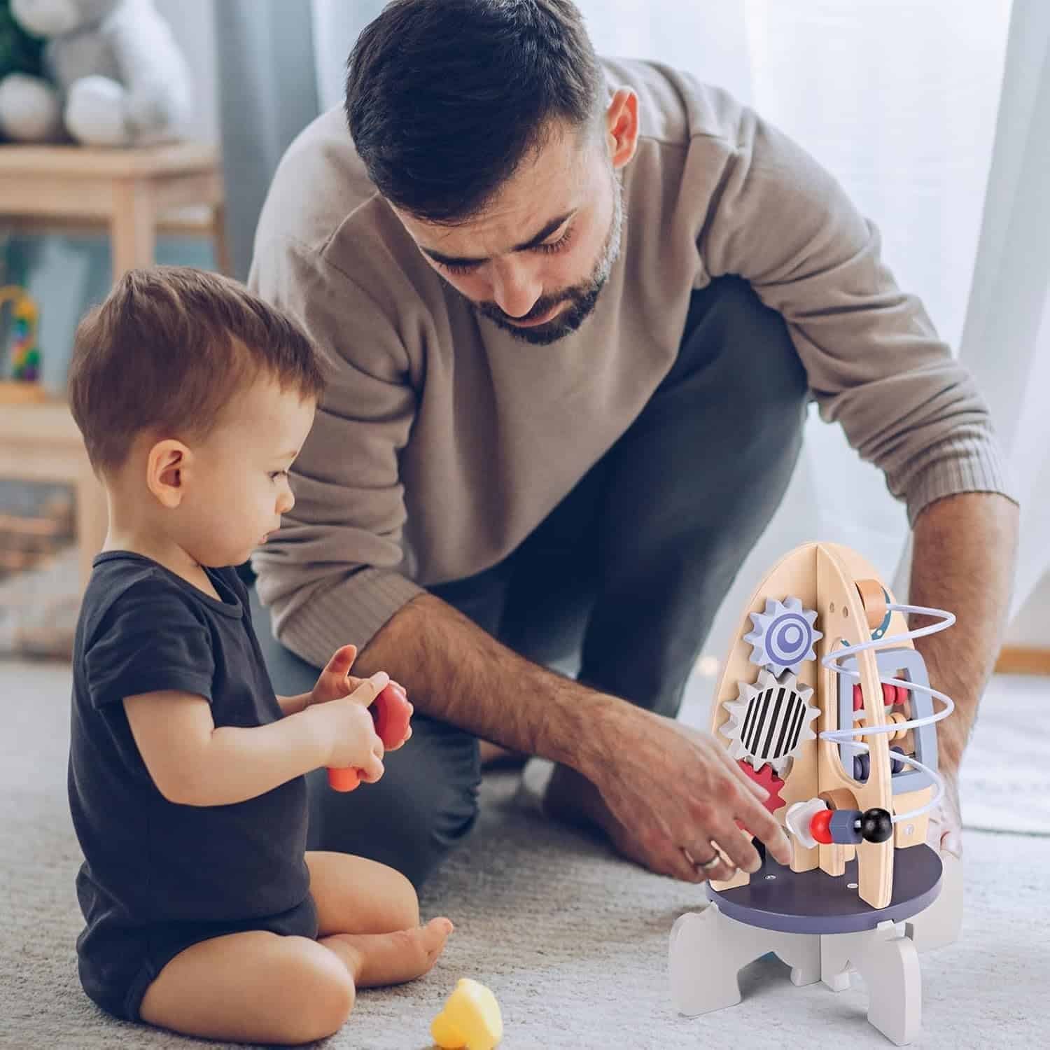 An adult and a young child sit on the floor together, interacting with a wooden rocket-shaped activity toy.
