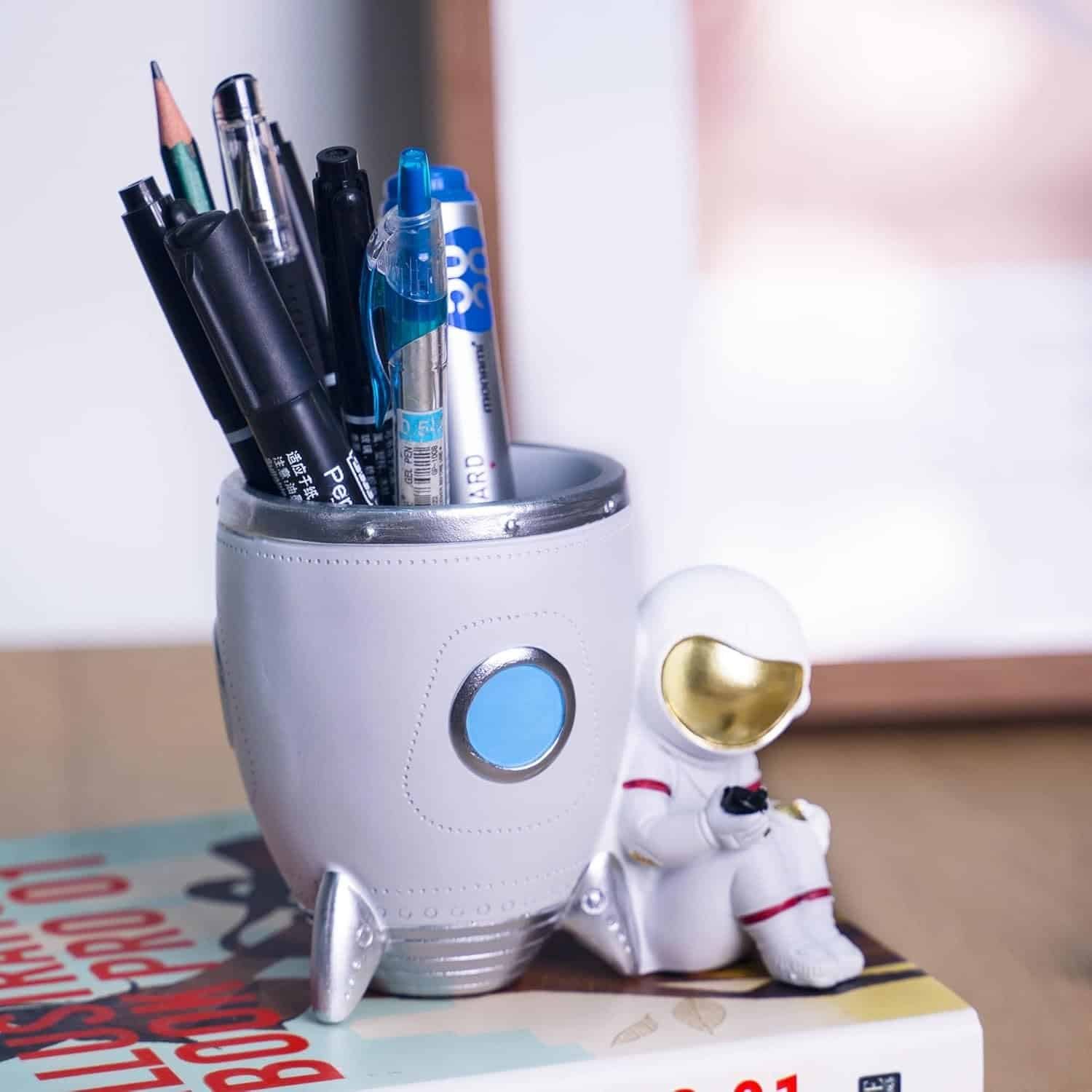 A rocket-shaped pen holder with pens and markers inside, next to a small astronaut figurine, sits on top of a closed book on a wooden surface.