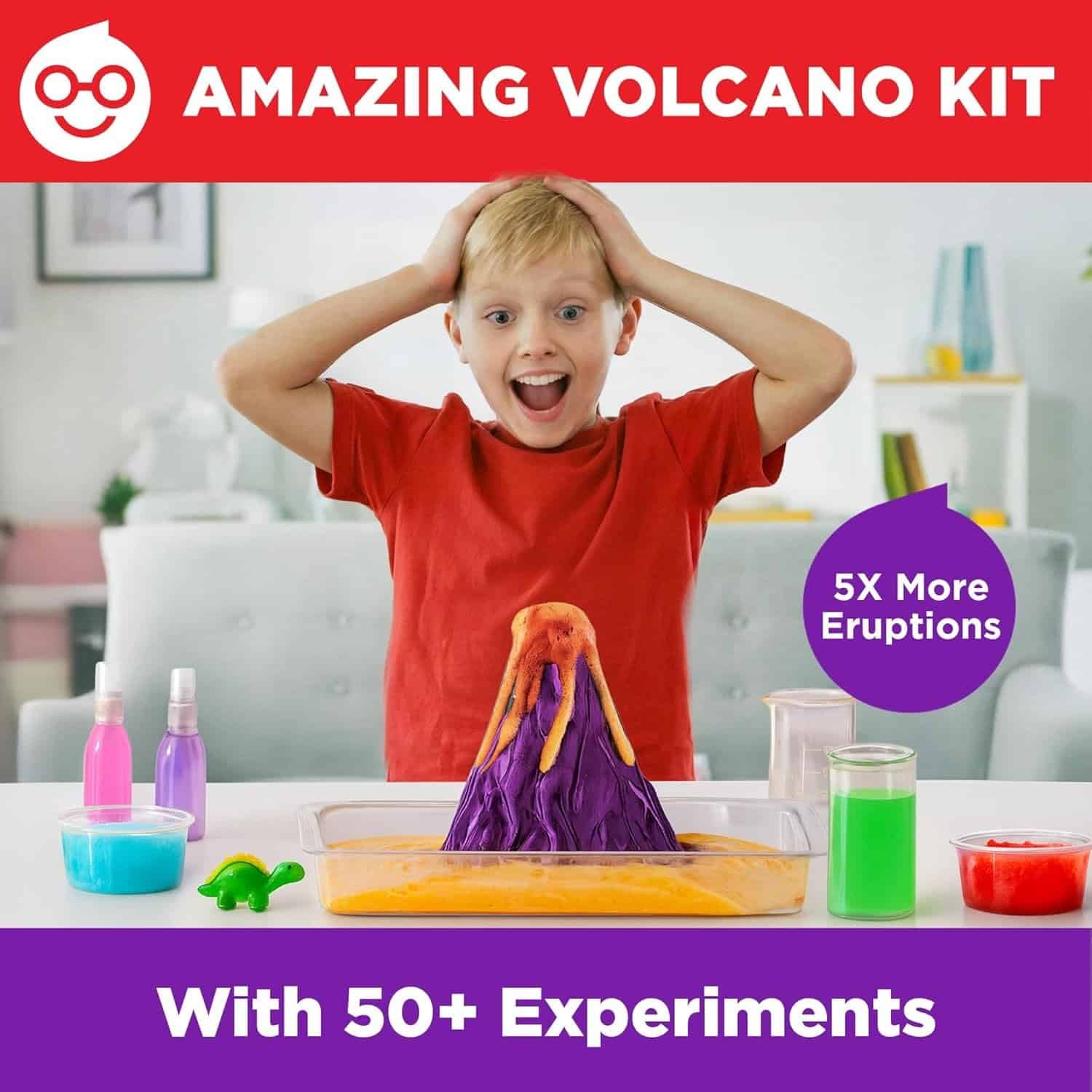 A young boy looks excited as a toy volcano erupts with orange foam. Science experiment bottles and jars are on the table. Text promotes an "Amazing Volcano Kit" with 50+ experiments.