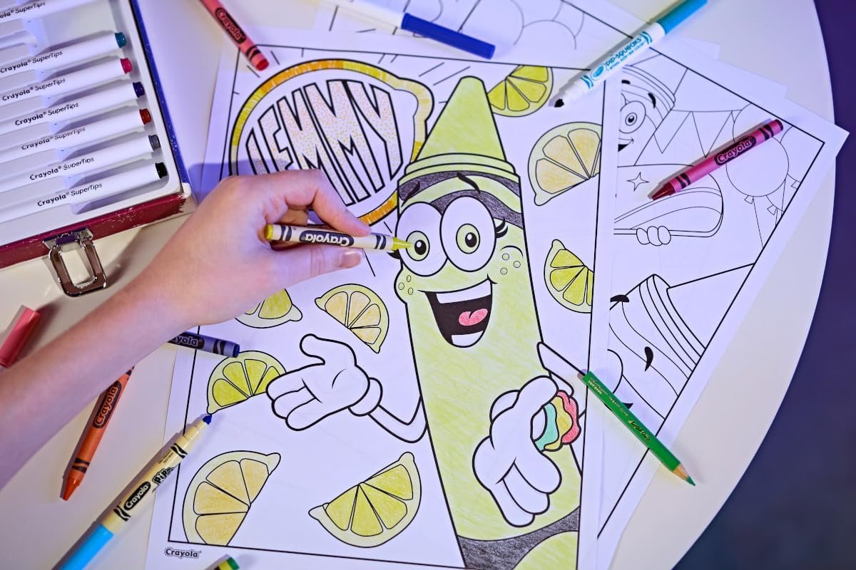 A hand colors a cartoon lemon character named "Lemmy" with yellow crayon on a coloring page, surrounded by other crayons and coloring sheets on a white table.