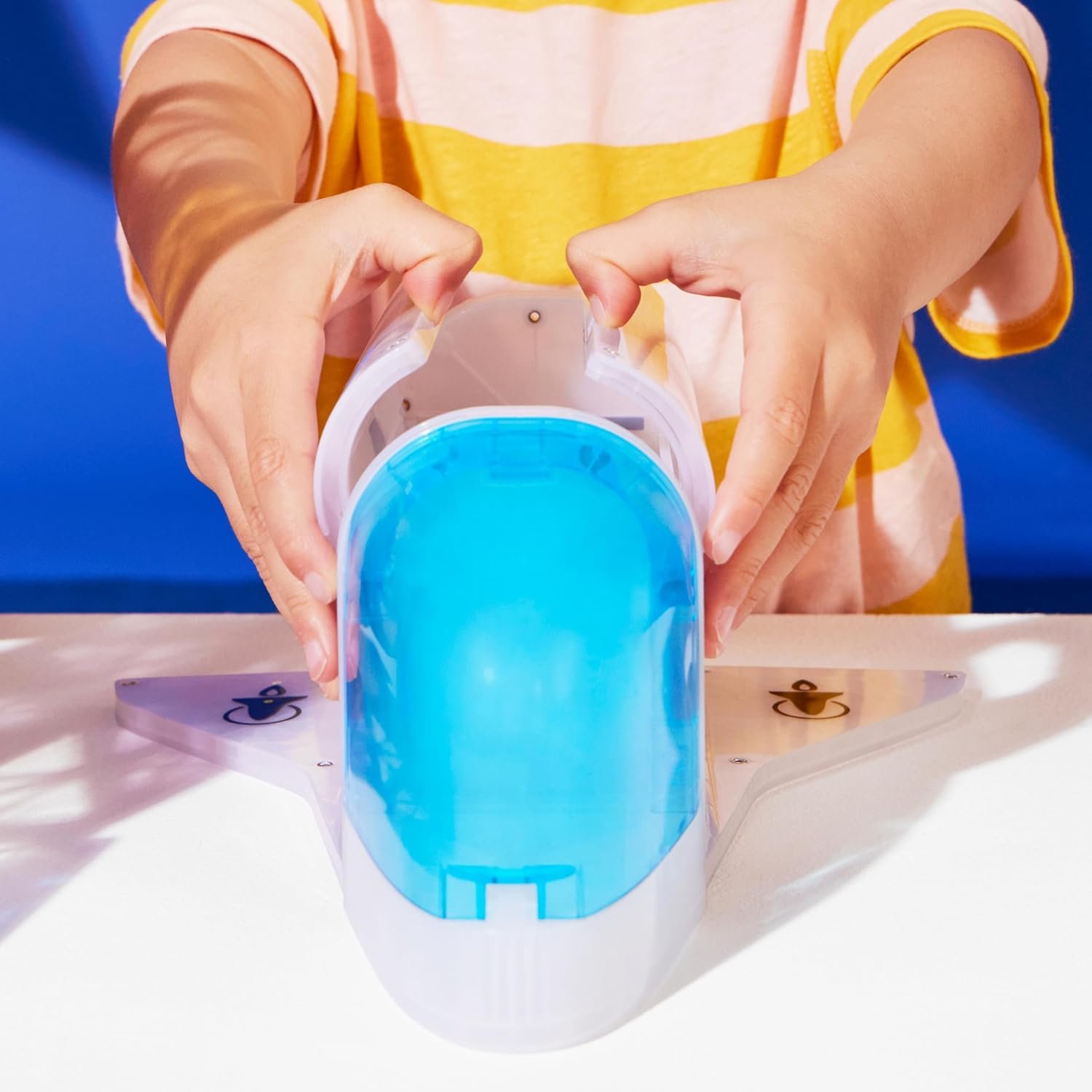 A child in a yellow striped shirt assembles or plays with a white plastic toy shaped like an airplane with a blue cockpit on a white table.