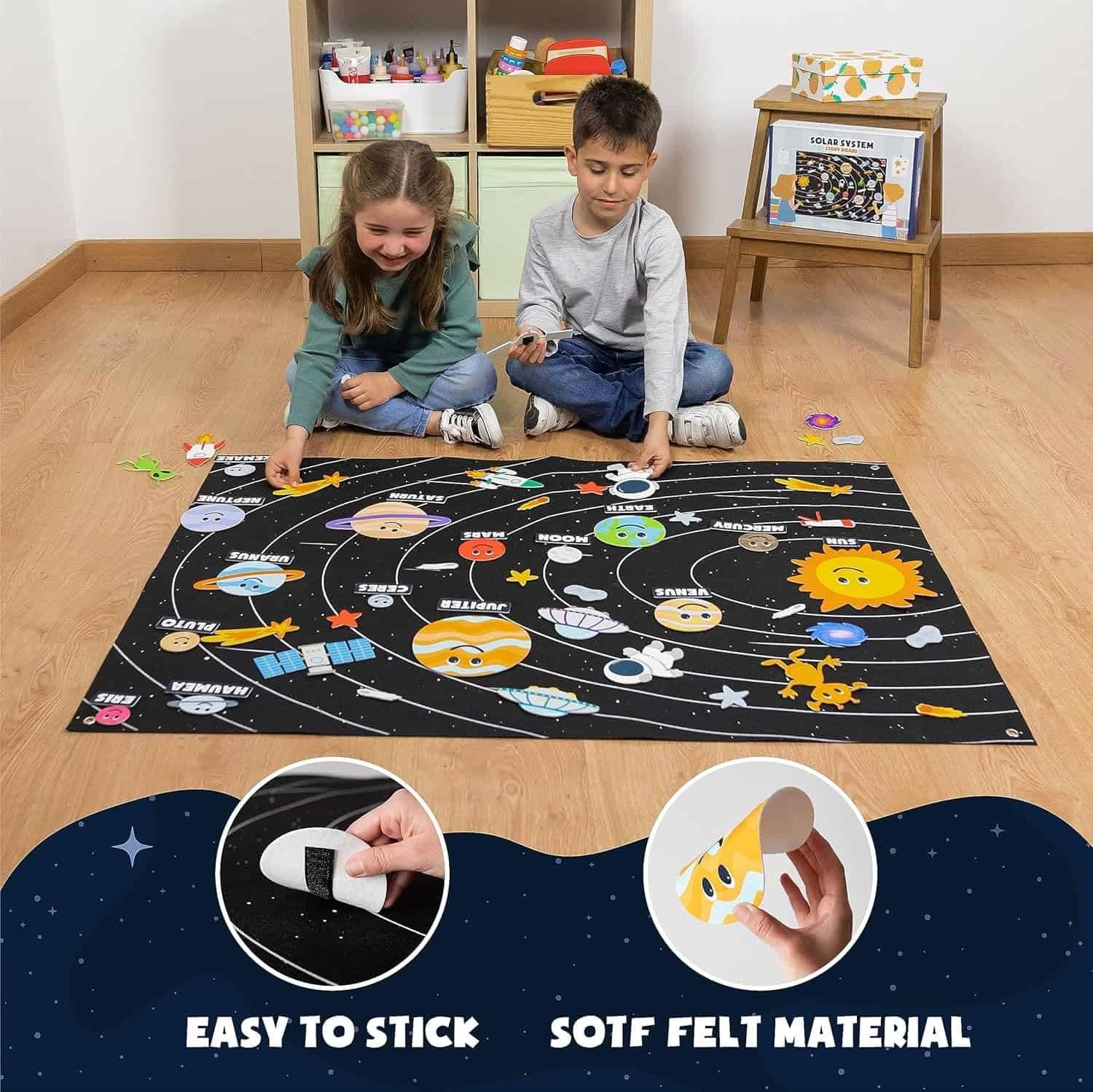 Two children sit on the floor playing with a solar system-themed felt board, attaching planets and space objects, with shelves of toys and books in the background.