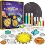 A National Geographic stained glass craft kit featuring planet designs, glow-in-the-dark paints, a solar system background mat, suction cups, ribbon, and crafting tools.