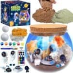 Space exploration-themed terrarium kit with astronaut figurines, UFOs, paints, moss, sand, remote control light, and tools displayed around a decorated glass container and box.