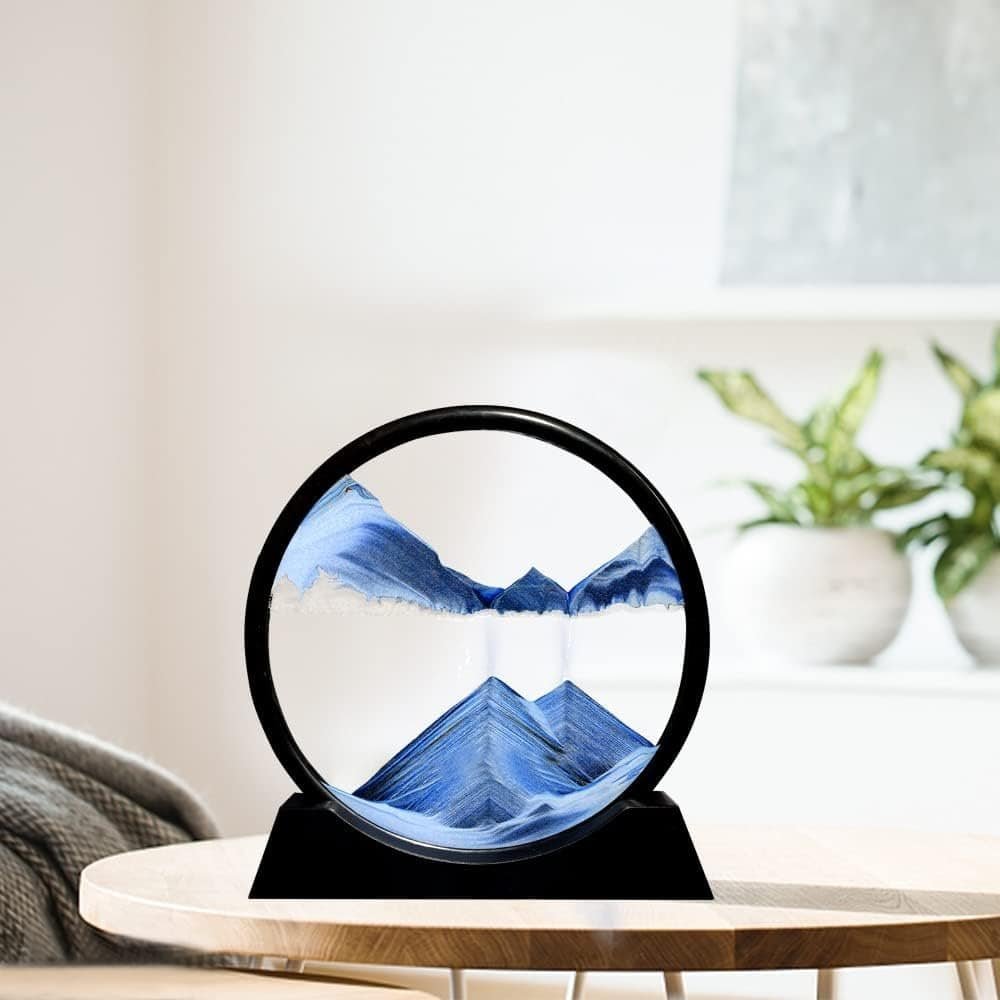 A circular sand art sculpture with blue and white sand forming mountain-like patterns sits on a wooden table in a bright, modern room.