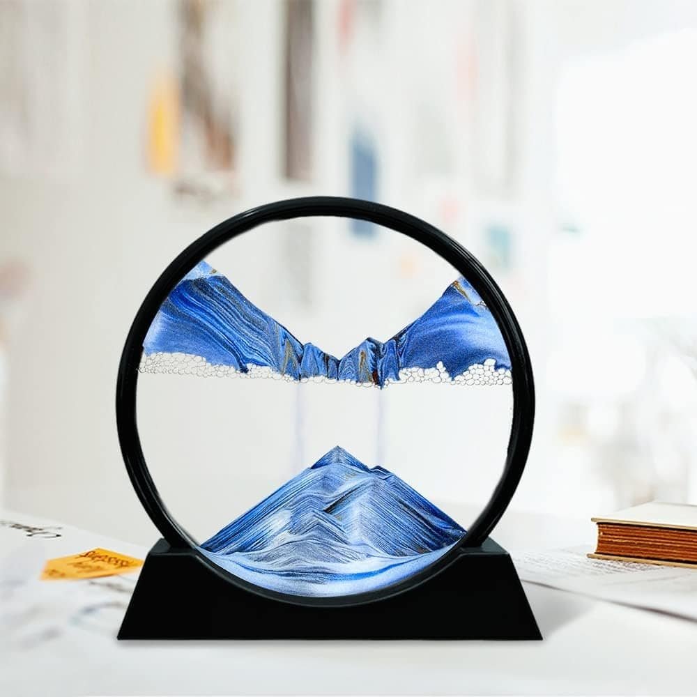 A circular sand art sculpture on a black stand displays blue and white sand forming mountain-like patterns, set on a white surface with blurred background.