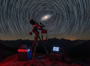 A telescope and computer equipment set on a rocky surface under a night sky time-lapse, capturing circular star trails centered around the North Star—a captivating scene perfect for astronomy enthusiasts.