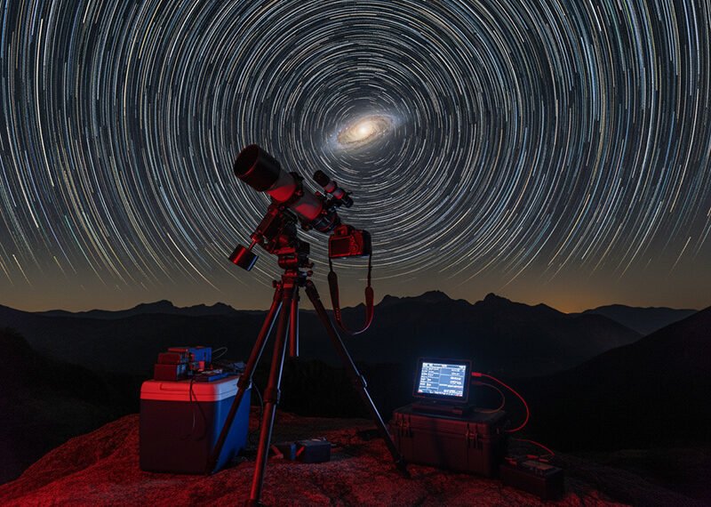 A telescope and computer equipment set on a rocky surface under a night sky time-lapse, capturing circular star trails centered around the North Star—a captivating scene perfect for astronomy enthusiasts.
