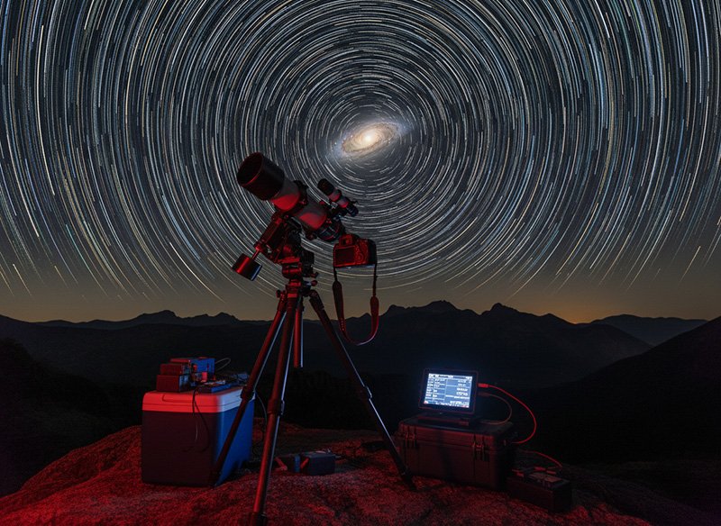 A telescope and computer equipment set on a rocky surface under a night sky time-lapse, capturing circular star trails centered around the North Star—a captivating scene perfect for astronomy enthusiasts.
