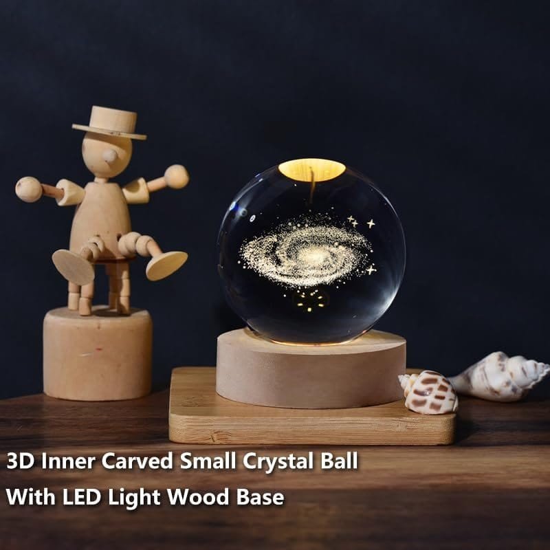 A glass crystal ball with a carved galaxy inside sits on a lit wooden base, next to a wooden figure and seashells on a wooden surface. Text reads: "3D Inner Carved Small Crystal Ball With LED Light Wood Base.