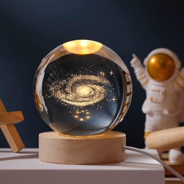 A glass sphere with a galaxy design inside sits on a wooden base, softly lit from above, with a small astronaut figurine in the background.