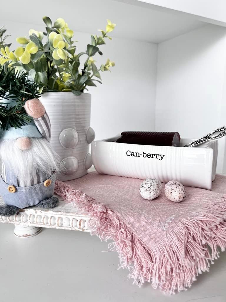 A small ceramic gnome, a potted plant, a white dish labeled "Can-berry" with cranberry sauce, two speckled candy eggs, and a pink fringed cloth arranged on a white shelf.