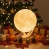 A hand-shaped lamp holding a glowing moon sphere sits on a table with pumpkins, pinecones, leaves, and wrapped gifts, in front of a decorated Christmas tree with lights.