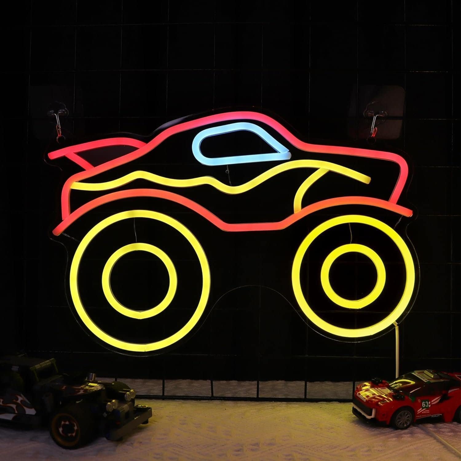 Neon sign in the shape of a monster truck is lit up in yellow, red, and blue; two toy cars are placed below the sign.