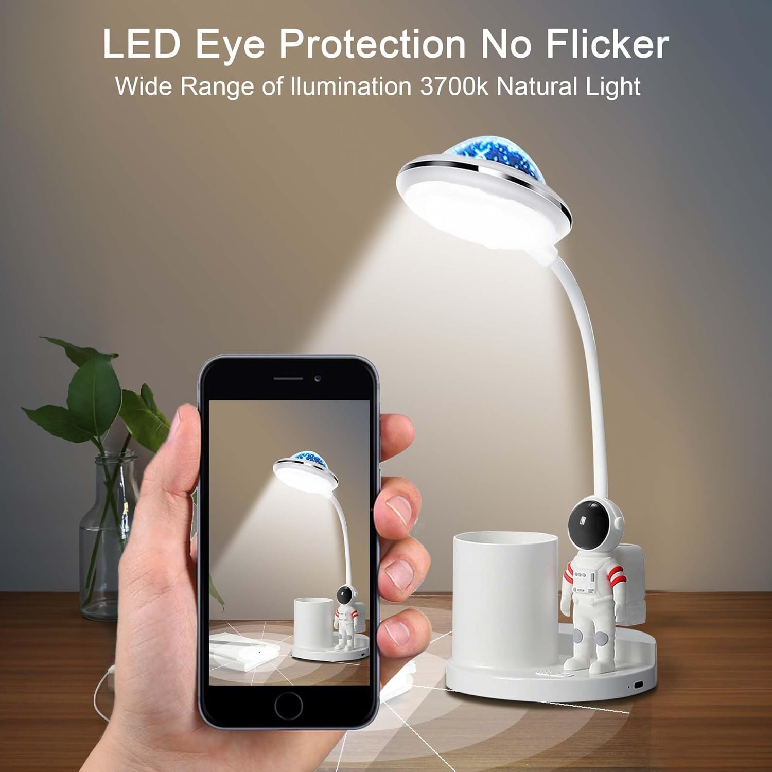 A person holds a smartphone taking a photo of a white LED desk lamp shaped like an astronaut with a cup holder on a wooden surface under soft lighting.