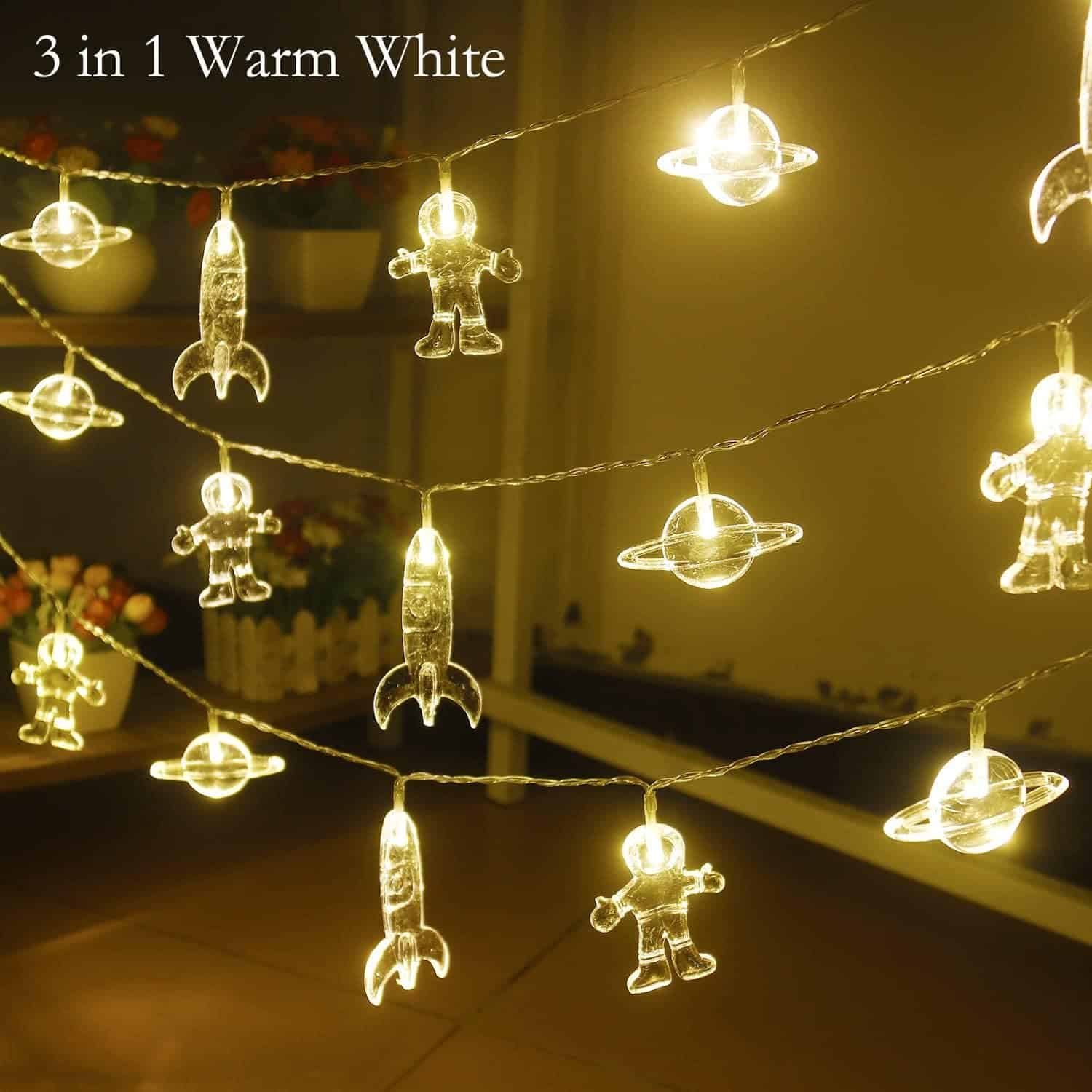 String lights with glowing astronaut, rocket, and planet shapes in warm white color, hanging indoors with a decorative background. Text reads "3 in 1 Warm White.