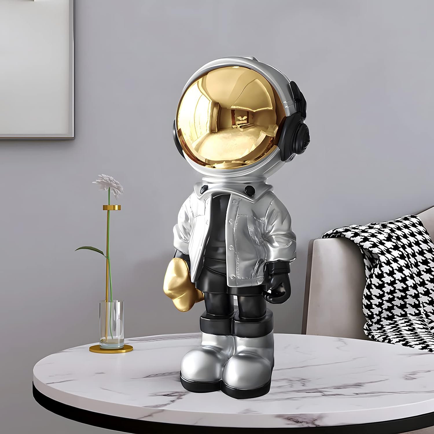 A small astronaut figurine with a gold helmet and silver outfit stands on a round marble table next to a vase with a single white flower in a modern living room.