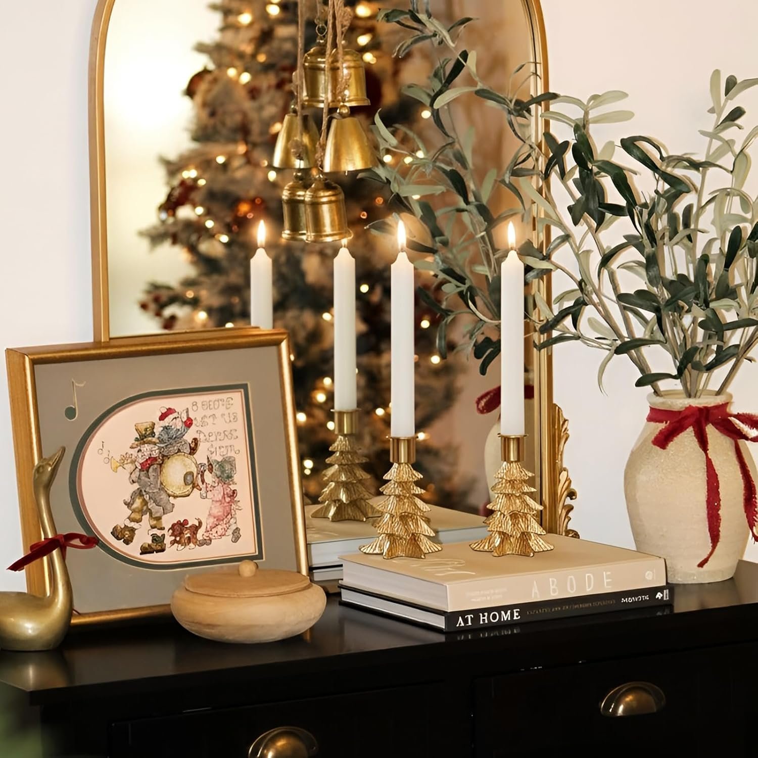A holiday-themed table display featuring three lit candles in gold tree-shaped holders, a vase with greenery, a framed illustration, books, and a gold bell garland, with a mirror reflecting a Christmas tree.