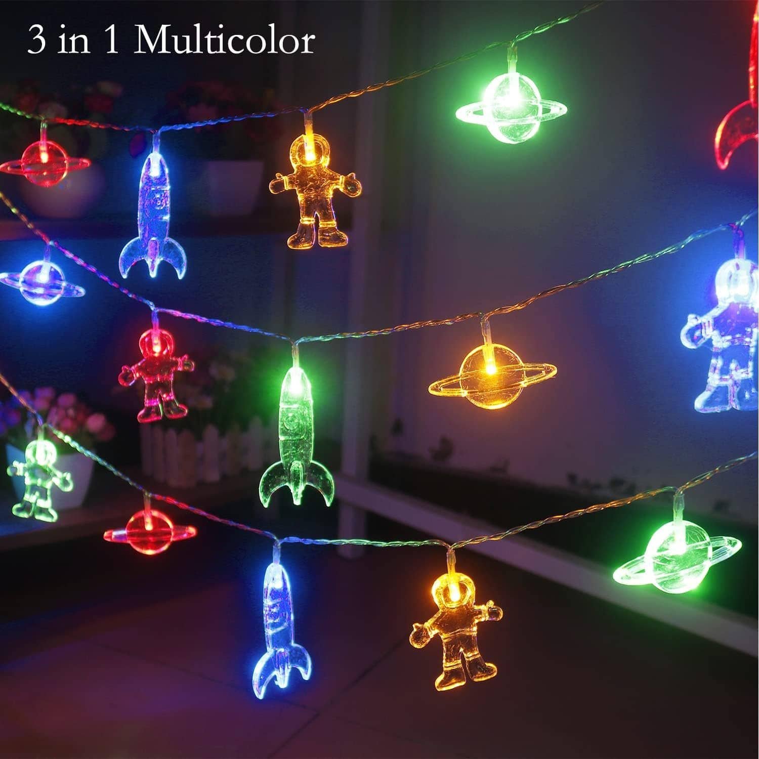 Multicolor string lights featuring astronauts, rockets, and planets are hanging outdoors at night, glowing in red, green, yellow, and blue. Text reads "3 in 1 Multicolor.