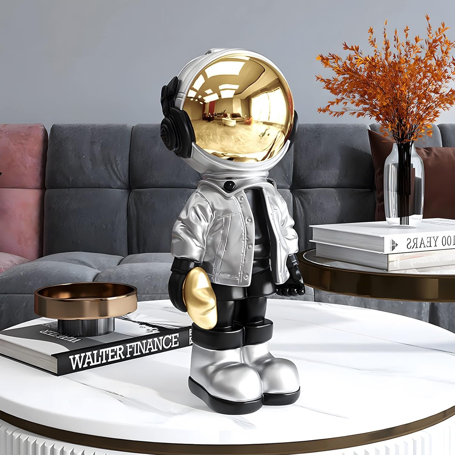 A metallic astronaut figurine with a gold helmet stands on a white coffee table, next to books and a decorative vase in a modern living room setting.