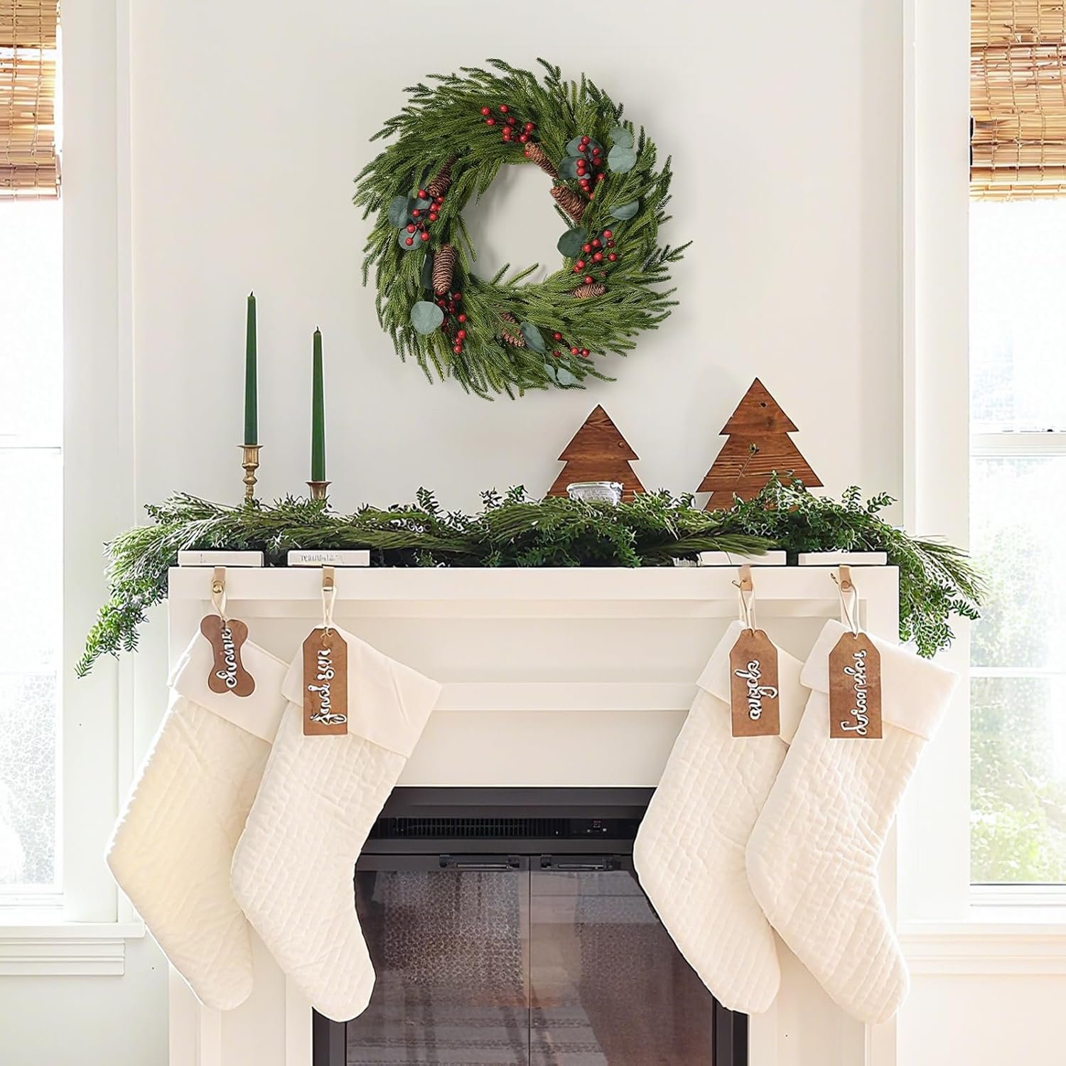 A white fireplace mantel decorated with greenery, two green candles, wooden trees, a wreath above, and four white stockings labeled with names hanging below.