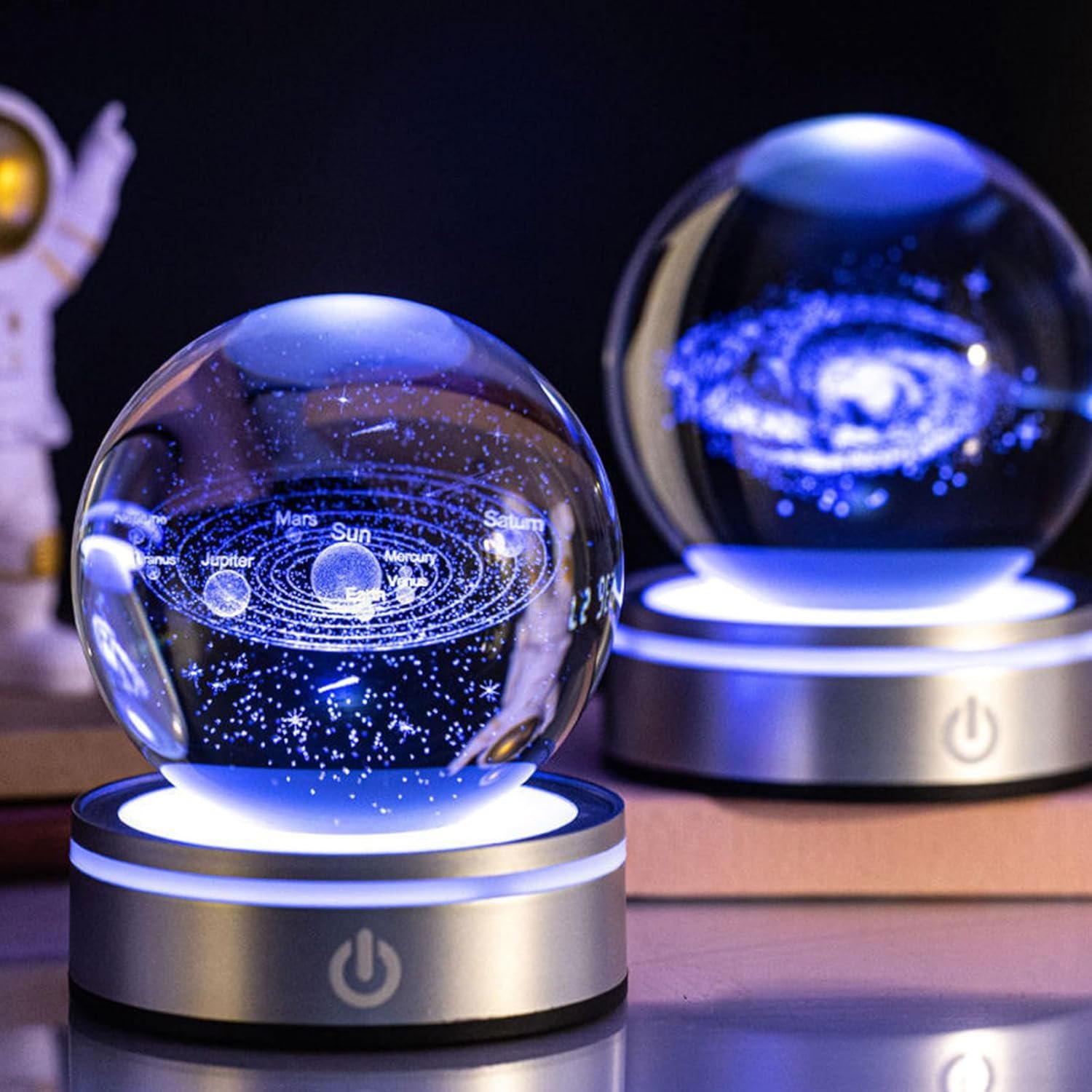 Two illuminated glass orbs on stands display detailed engravings of the solar system and a spiral galaxy, with an astronaut figurine blurred in the background.