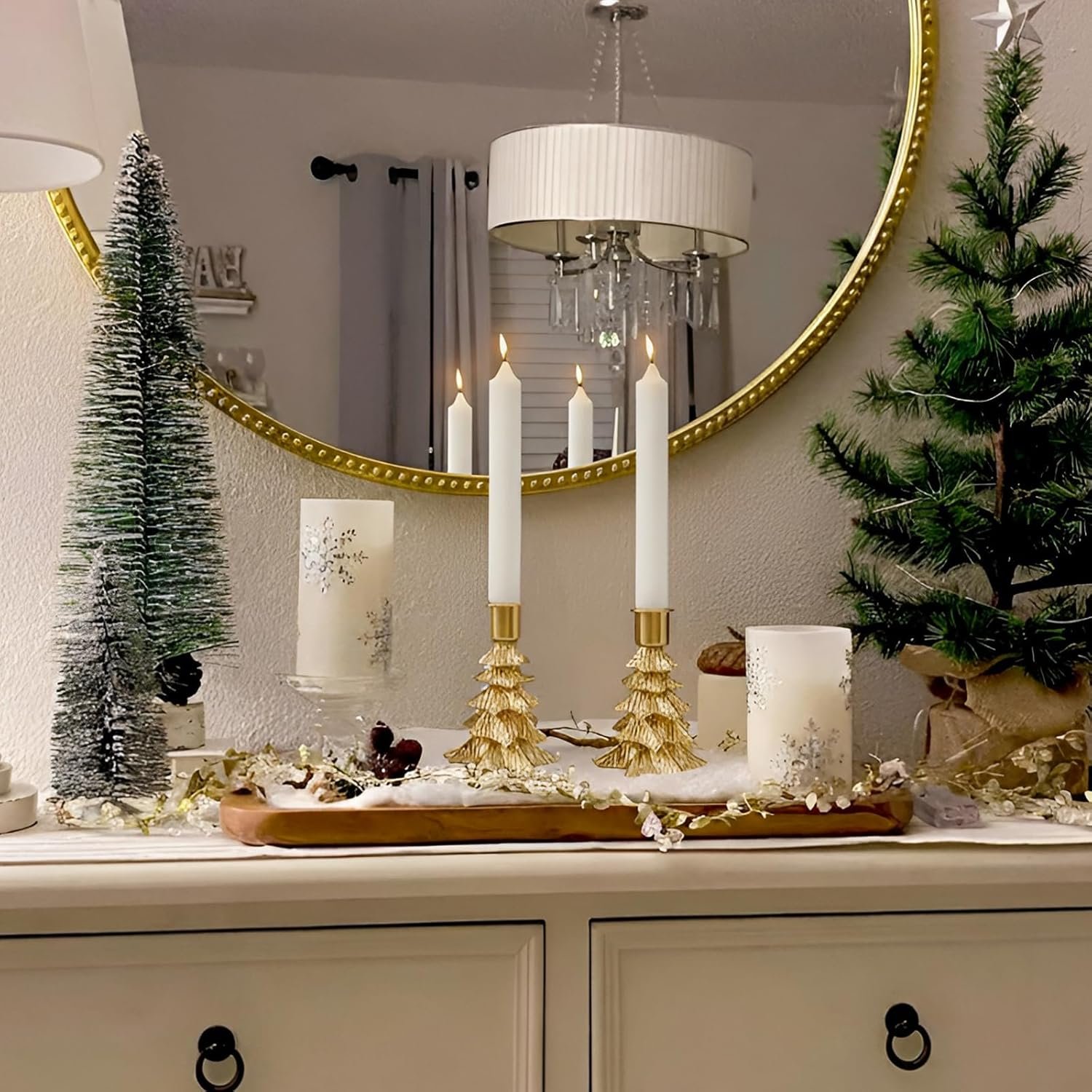 A decorated dresser with two white candles in gold holders, artificial Christmas trees, and holiday-themed candles, set in front of a round mirror.