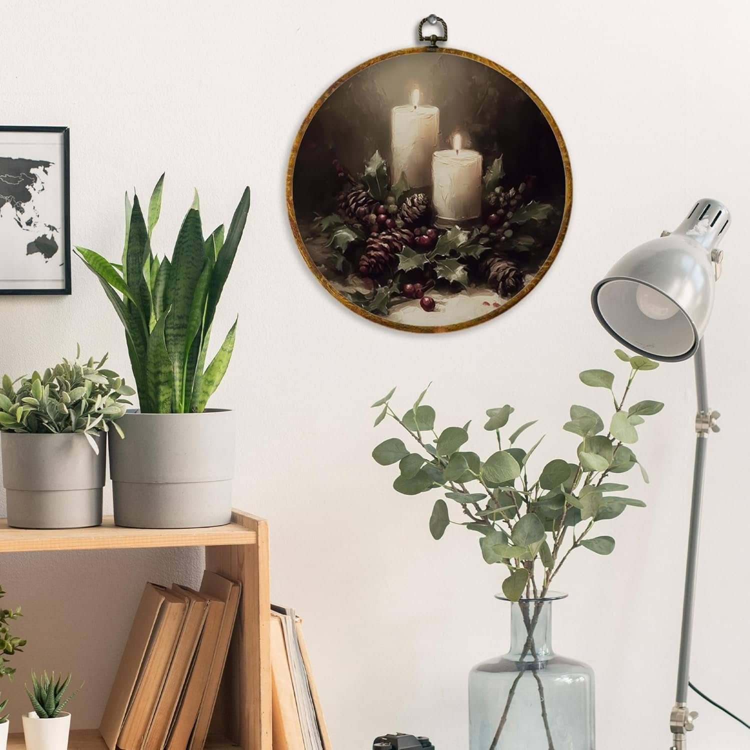 Wall art featuring two white candles, pinecones, and holly hangs above a shelf with potted plants, books, and a vase with leafy branches next to a silver desk lamp.