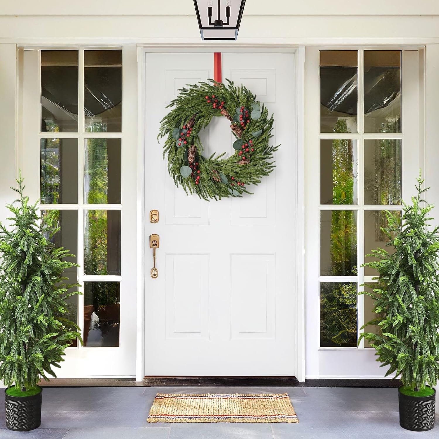 A white front door with a holiday wreath, flanked by two potted evergreen trees, and a small doormat on the porch.