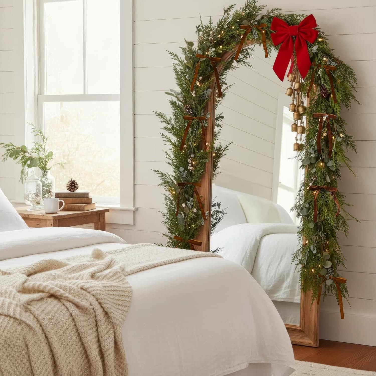 A large mirror in a bedroom is decorated with a garland of greenery, string lights, and a red bow. The bed has white bedding and a beige knit blanket.