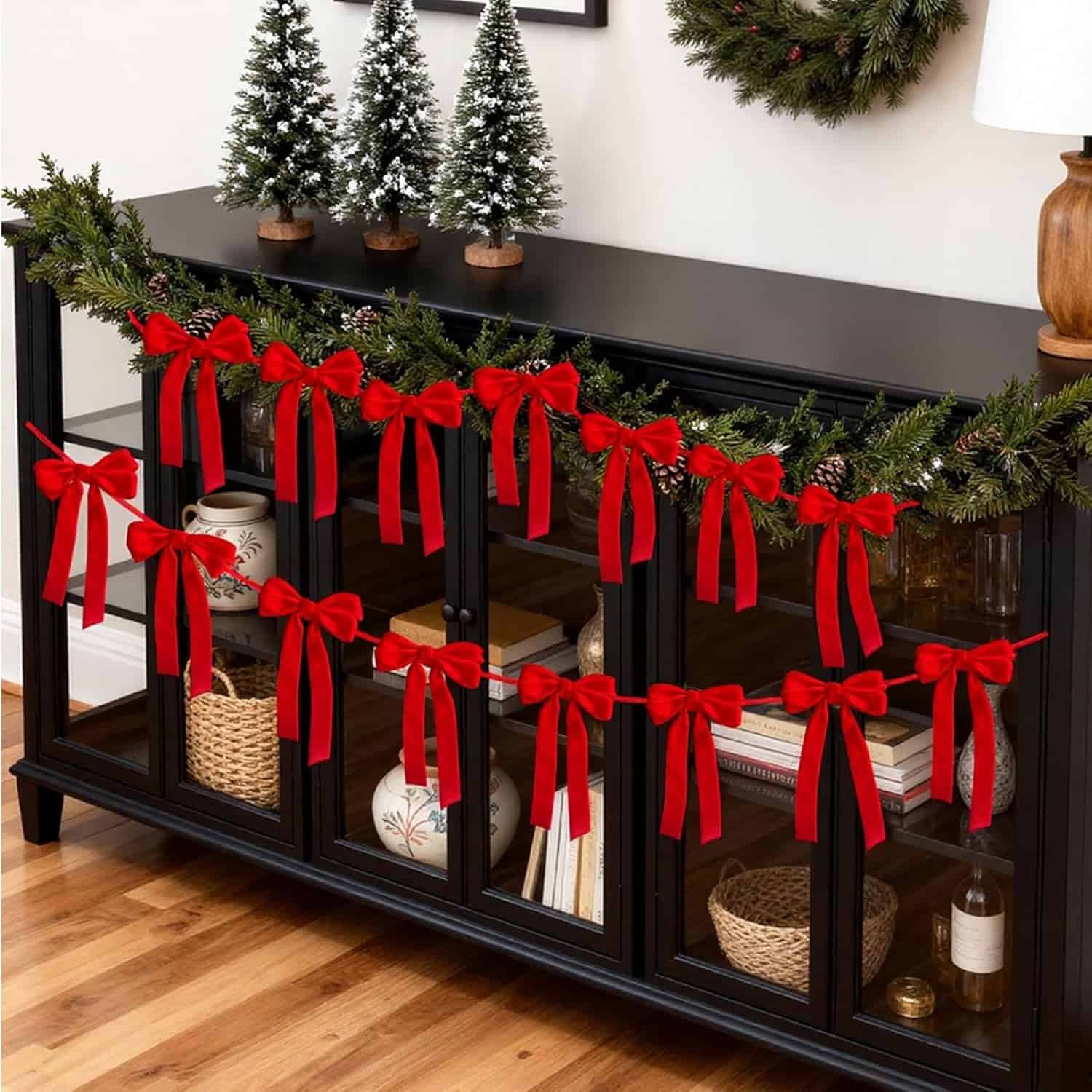 A black sideboard decorated with a garland and red bows, topped with three small Christmas trees and a wreath on the wall above.