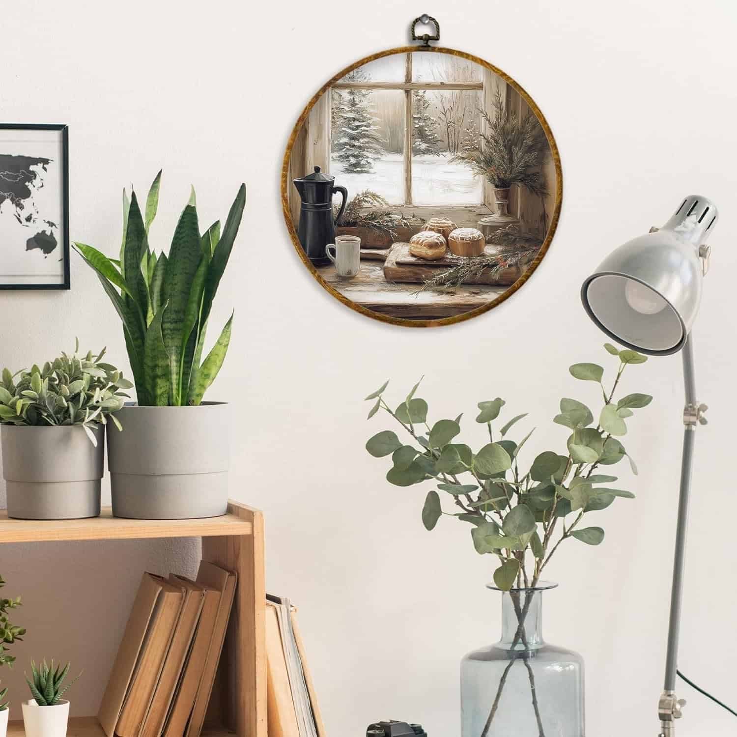 A room with houseplants, books, a metal desk lamp, and a round wall art showing a winter window scene with coffee and pastries.